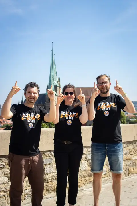 Drei Fans in „Ich bin ein Löwe“-Shirts zeigen nach oben, dahinter die Domspitzen